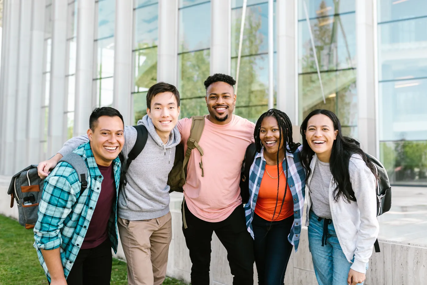 Five smiling college students with backpacks standing arm in arm outside a campus building